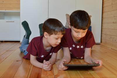 Bild vergrößern: Zwei Jungen in dunkelroten Shirts mit weißen Sternen mit kurzen dunklen Haaren liegen auf dem Holzfußboden und schauen auf ein Tablet.
